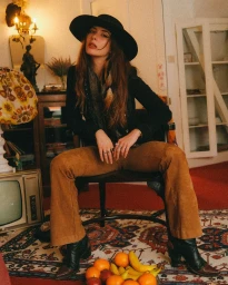 Woman in a black wide-brim hat and suede trousers seated on a chair indoors, photographed by Bob Sala