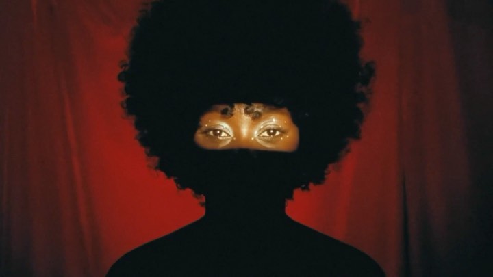 Dark portrait of a Black woman with an afro and gold eyeshadow, partially veiled by a black fabric against a blurred red background.