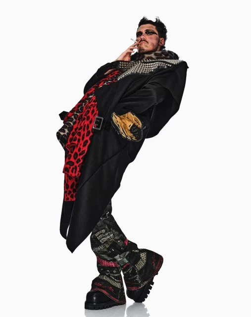 A slender man with dark eye makeup stands indoors in a studded black coat, animal print scarf, patterned pants, and platform boots smoking a cigarette.