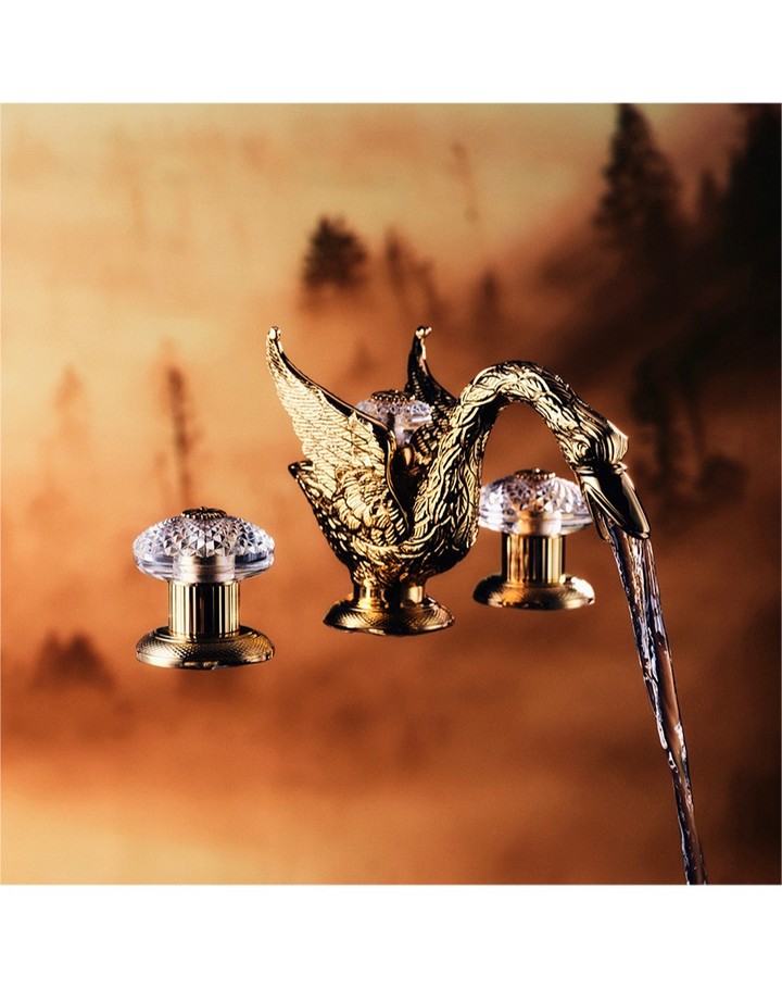Close-up of a gold swan faucet with crystal handles, water flowing, set against a blurry background of trees and golden light.