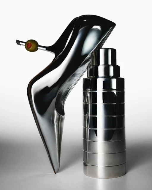 Stylish still life: a silver stiletto shoe with an olive garnish leans against a sleek, metallic cocktail shaker on a white surface.