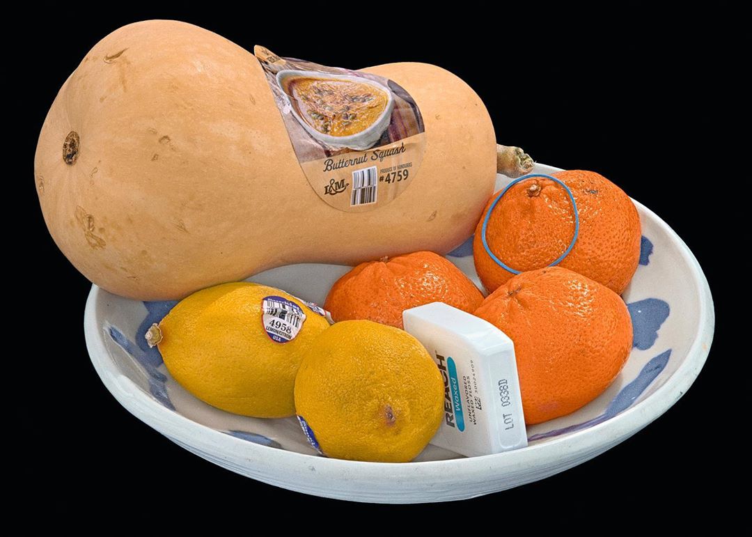 Still life shot of a butternut squash, lemons, and oranges in a white bowl against a black backdrop, showcasing vibrant colors and textures.