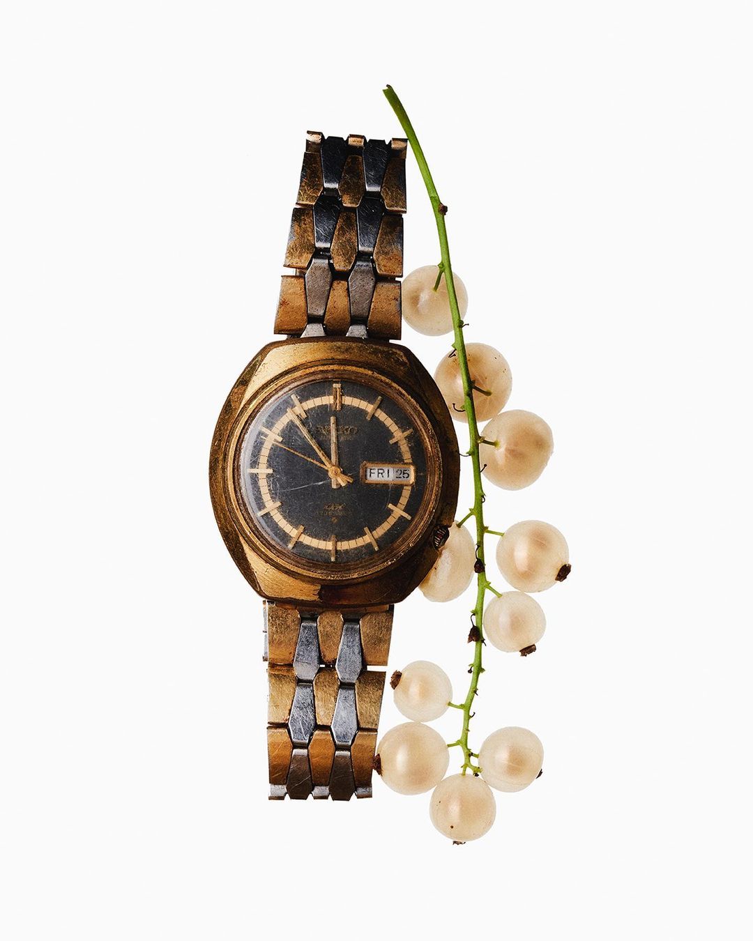 Wristwatch on a metal band next to a sprig of white currant berries on a white backdrop in a still life product shot.