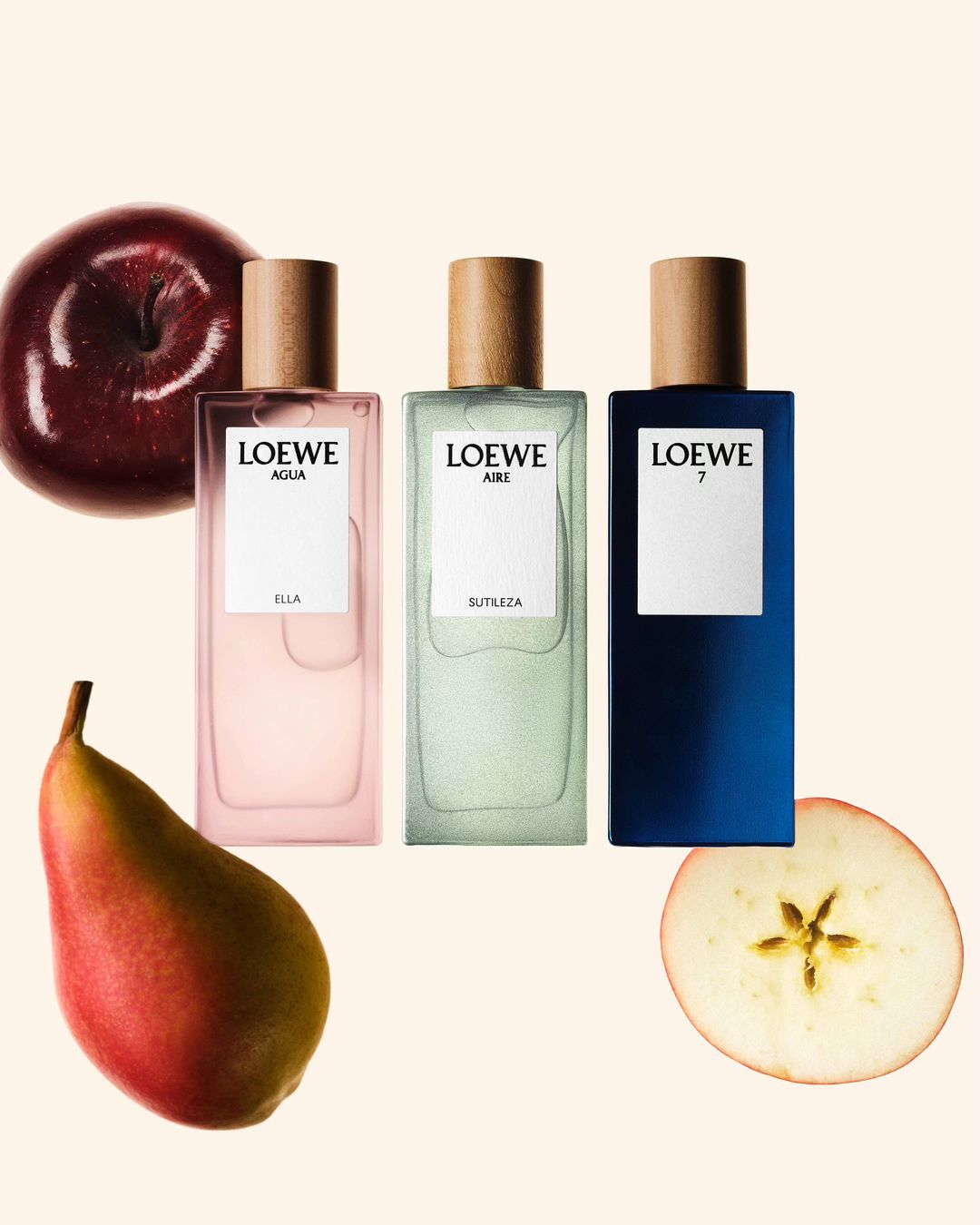 Three Loewe perfume bottles are arranged with a sliced apple, pear, and whole apple against a light tan background in a studio setting.
