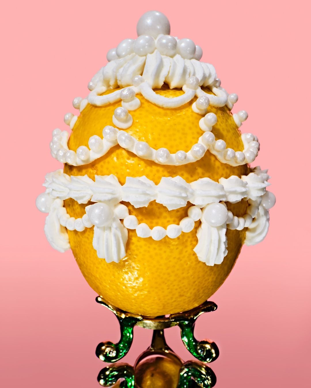 Ornate yellow lemon decorated with white icing and beads on a gold and green stand against a pink background in a close-up shot.