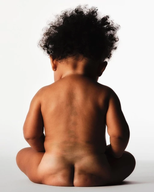 Infant with dark curly hair sits with back facing camera against white backdrop in studio lighting. Child portrait, baby photography.
