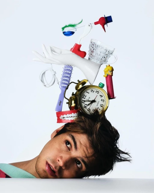 Man with whimsical items on his head including a clock, false teeth, and a hair brush against a clean white backdrop in a studio setting.