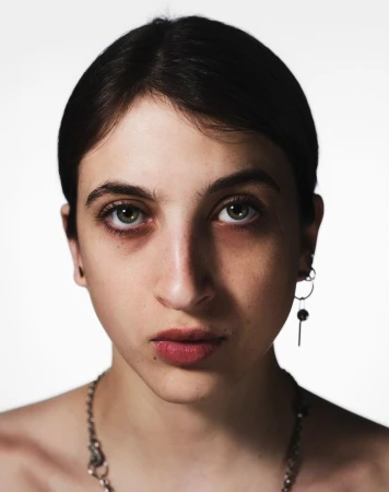 Close-up portrait of a young adult female with fair skin, dark hair, green eyes, and visible jewelry against a white background.