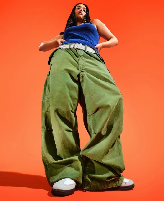 Woman in a blue tank top and baggy green pants stands against an orange background, captured in a low-angle shot emphasizing her figure.