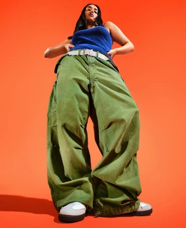 Woman in a blue tank top and baggy green pants stands against an orange background, captured in a low-angle shot emphasizing her figure.
