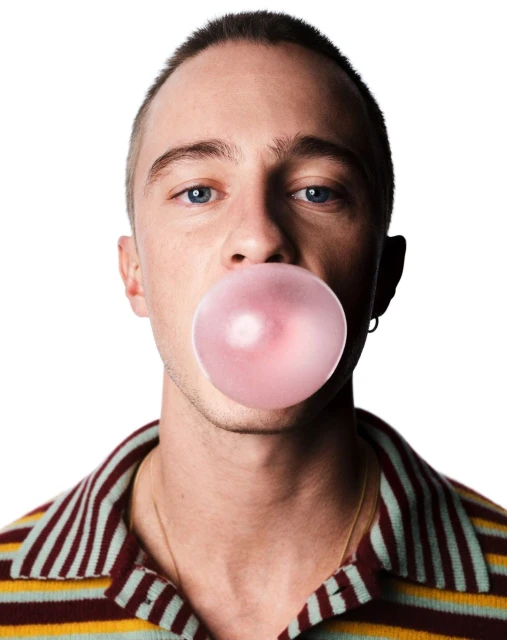 A young adult male with light skin blowing a pink bubblegum bubble, wearing a striped shirt against a stark white studio backdrop.