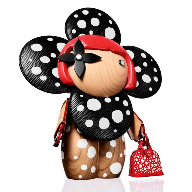 Whimsical wooden doll with red wig, black floral hair clip, and black polka-dot attire carrying a small red purse on white.