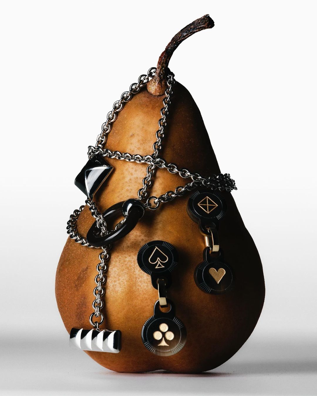 A pear is decorated with a silver chain and black and white charms shaped like playing card suits against a white background.
