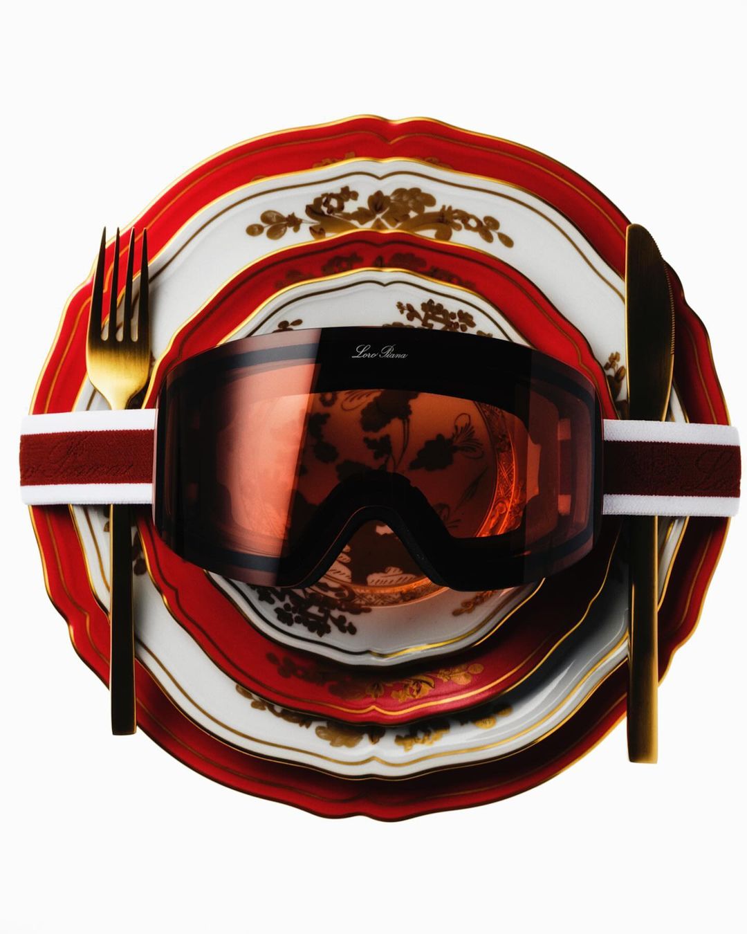 Ski goggles on a stack of decorative plates with a fork and knife. Isolated studio shot with a white background.