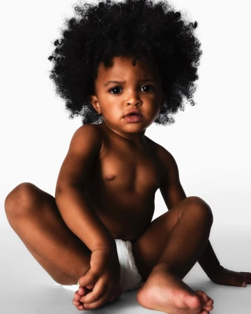 A young child with afro hair sits on a white background, wearing a diaper, looking at the camera with soft, diffused lighting.