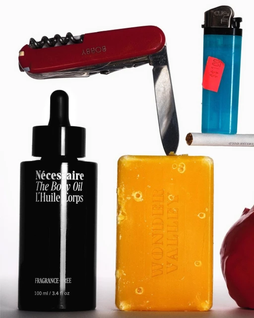A still life arrangement features a black bottle, red Swiss Army knife, yellow soap, blue lighter, cigarette, and an apple against white.