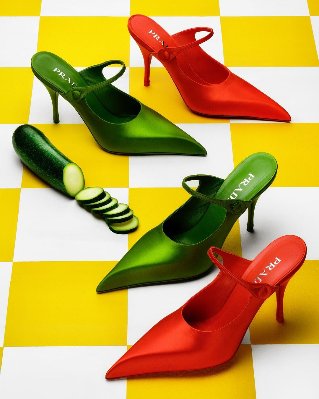 Still life of two pairs of women's heeled shoes, one green and one red, placed among zucchini on a yellow and white checkered surface.