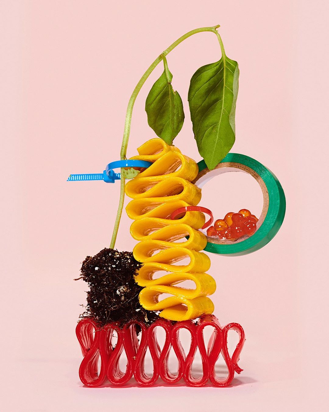 Creative still life composition on pink backdrop: yellow rolled pasta, red licorice, plant with green leaves, and green tape roll.