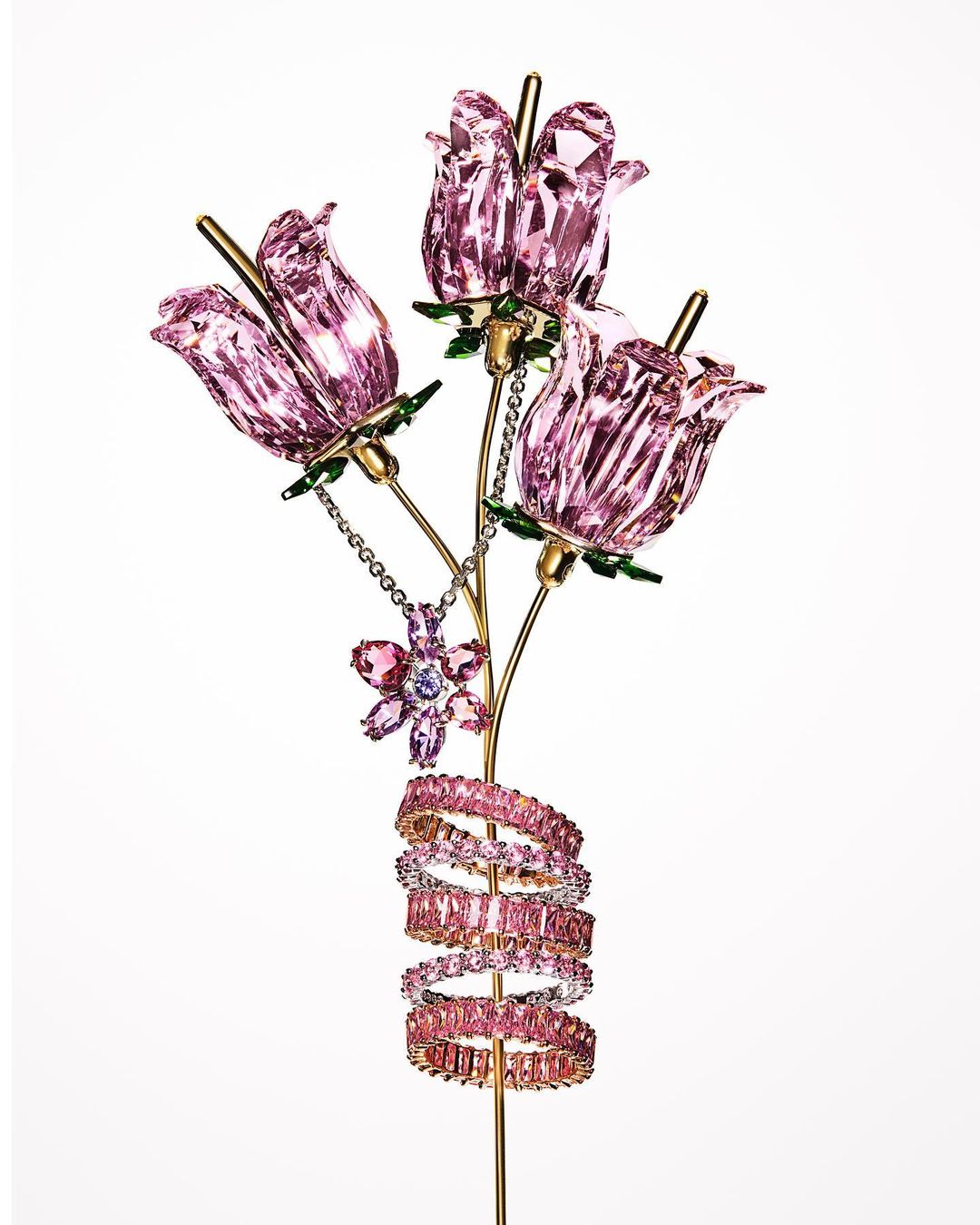 Crystal sculpture featuring three pink flower blossoms, a flower pendant, and pink gemstone rings wound around a golden stem, on white.