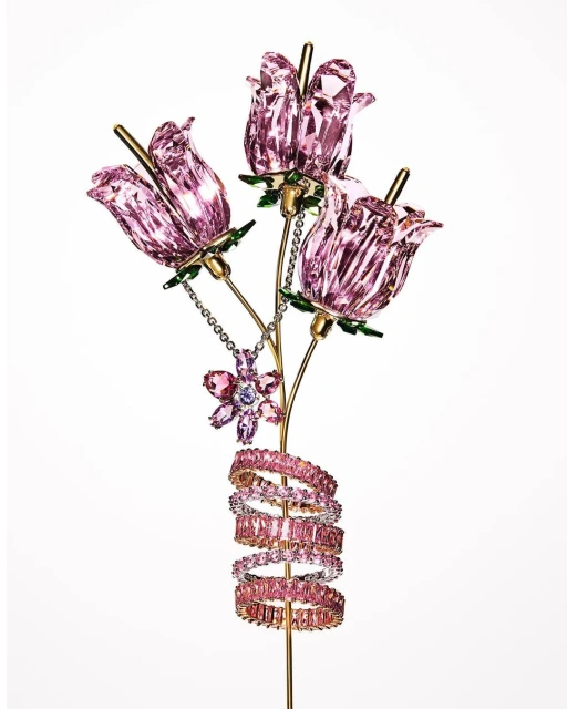 Crystal sculpture featuring three pink flower blossoms, a flower pendant, and pink gemstone rings wound around a golden stem, on white.