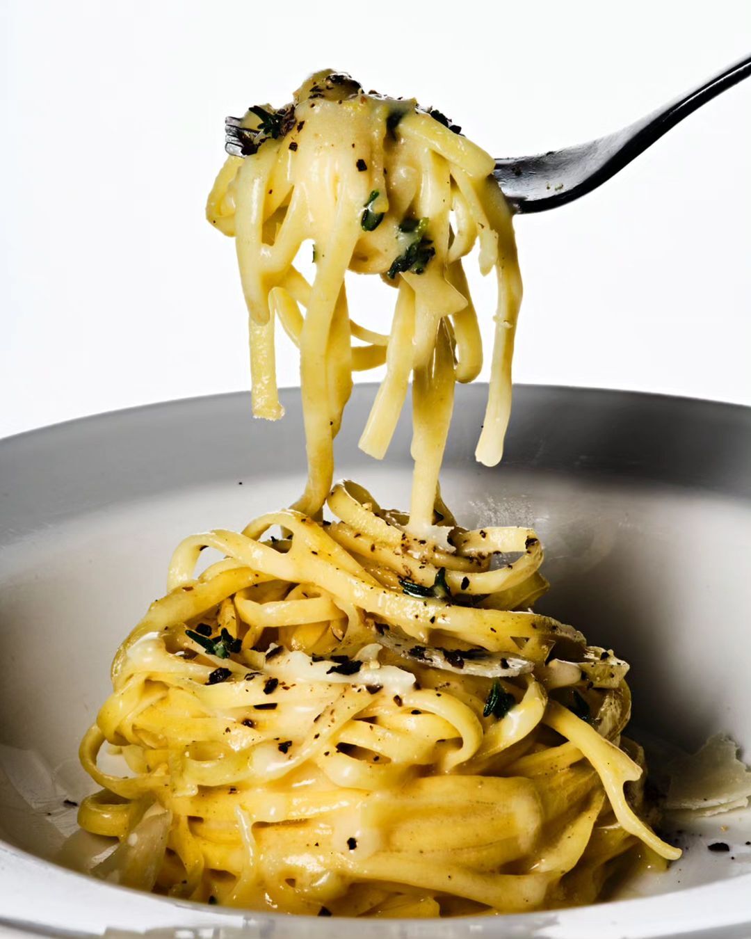 Close-up of creamy pasta with dark herbs on a fork and in a bowl against a white background in a brightly lit food shot.
