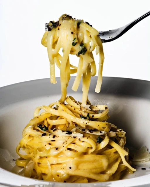 Close-up of creamy pasta with dark herbs on a fork and in a bowl against a white background in a brightly lit food shot.