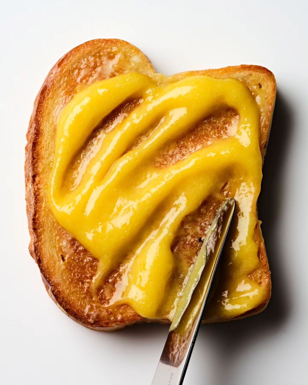 Overhead shot of toast covered in yellow spread, being applied with a silver butter knife on a plain white surface in bright lighting.