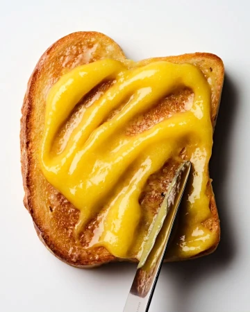 Overhead shot of toast covered in yellow spread, being applied with a silver butter knife on a plain white surface in bright lighting.