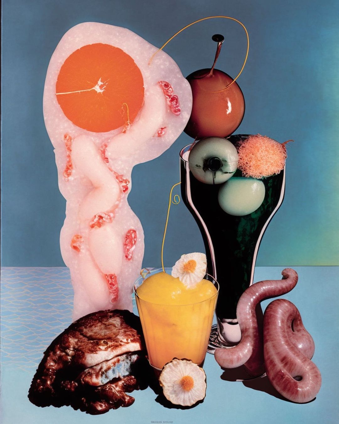 Still life composition features a surreal arrangement of food-like objects in vibrant colors on a light blue background.