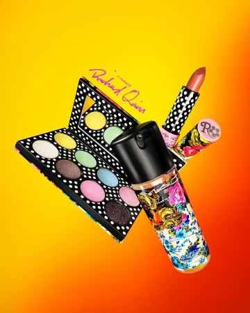 Still life shot of makeup products: eyeshadow palette, lipstick and a floral bottle, against a yellow and orange gradient background.
