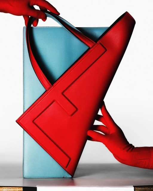 Fashion still life: Two disembodied hands holding a red leather handbag with a blue leather folio in front of a white background.