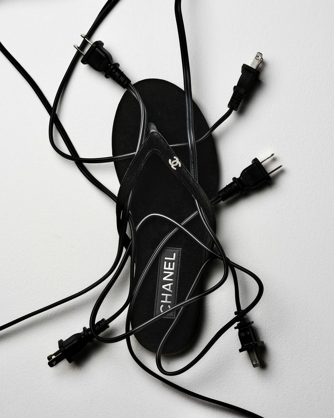 Black Chanel sandal surrounded by several electrical plugs with black cords on a white background in a fashion-style shot.
