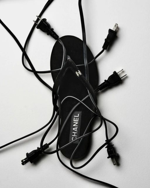 Black Chanel sandal surrounded by several electrical plugs with black cords on a white background in a fashion-style shot.