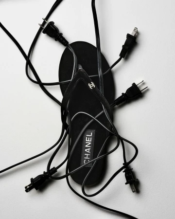 Black Chanel sandal surrounded by several electrical plugs with black cords on a white background in a fashion-style shot.