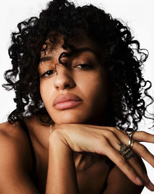 Close-up studio portrait of a young adult woman with dark, curly hair, wearing a spaghetti-strap top and rings, against a white backdrop.