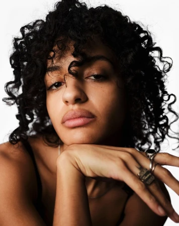 Close-up studio portrait of a young adult woman with dark, curly hair, wearing a spaghetti-strap top and rings, against a white backdrop.