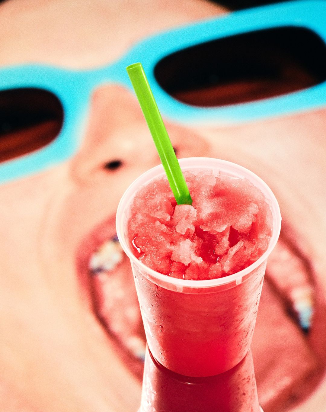 Close-up of child with sunglasses holding a red slush drink with green straw, bright and cheerful image with a summery, youthful vibe.