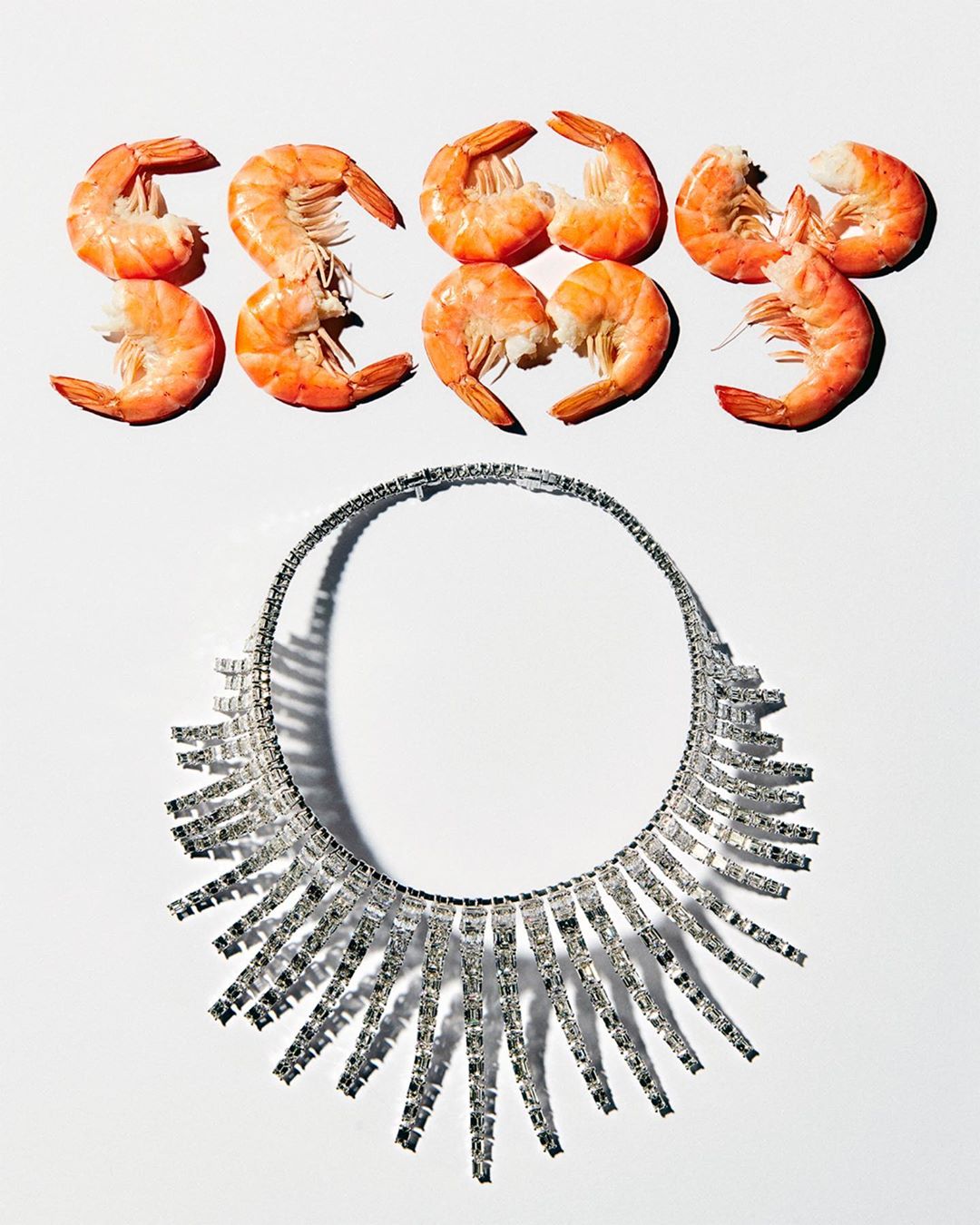 Overhead shot of cooked shrimp spelling 'SEXY' above a crystal statement necklace on a white background.