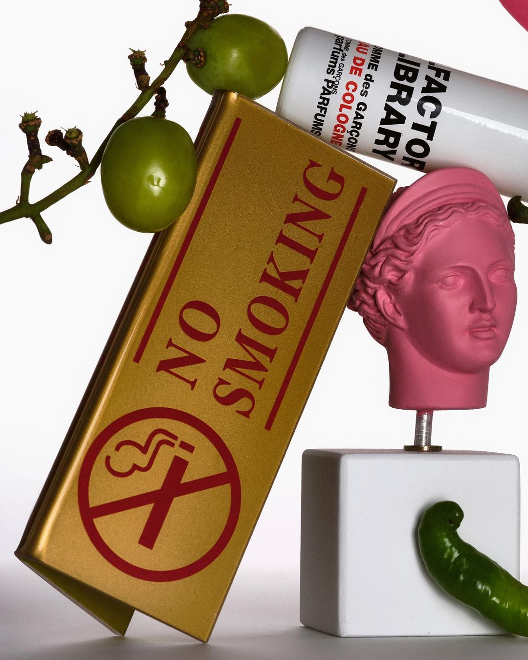 Still life composition with a pink head bust, grapes, a green pepper, perfume bottle, and a No Smoking sign against a white background.