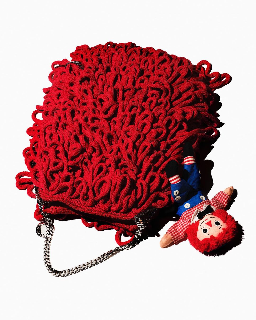 Red knit purse with chain strap laying next to a Raggedy Ann doll on a white background in a studio shot for fashion.