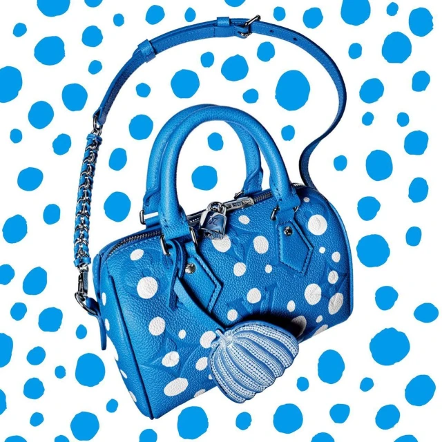Elevated eye-level studio shot of a blue Louis Vuitton handbag with white polka dots, complete with a matching charm, against a white polka-dot background.
