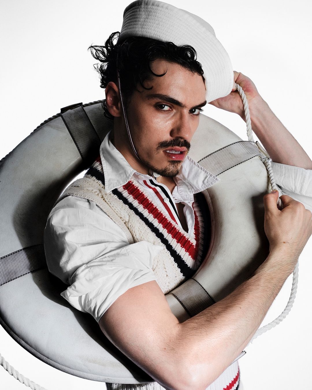 Portrait of a man with a mustache wearing a sailor's hat and a white and red cricket sweater, posing with a life preserver.