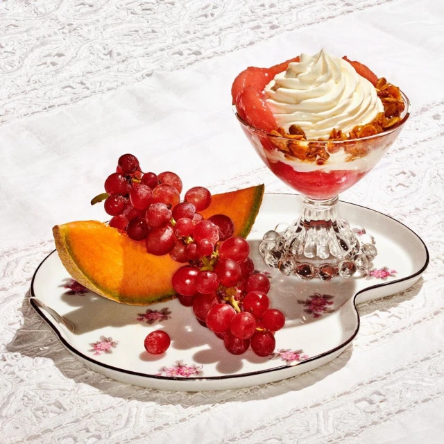 Close-up of a dessert featuring a parfait with whipped cream and fresh fruit, including melon slices and red grapes, presented on a floral plate.