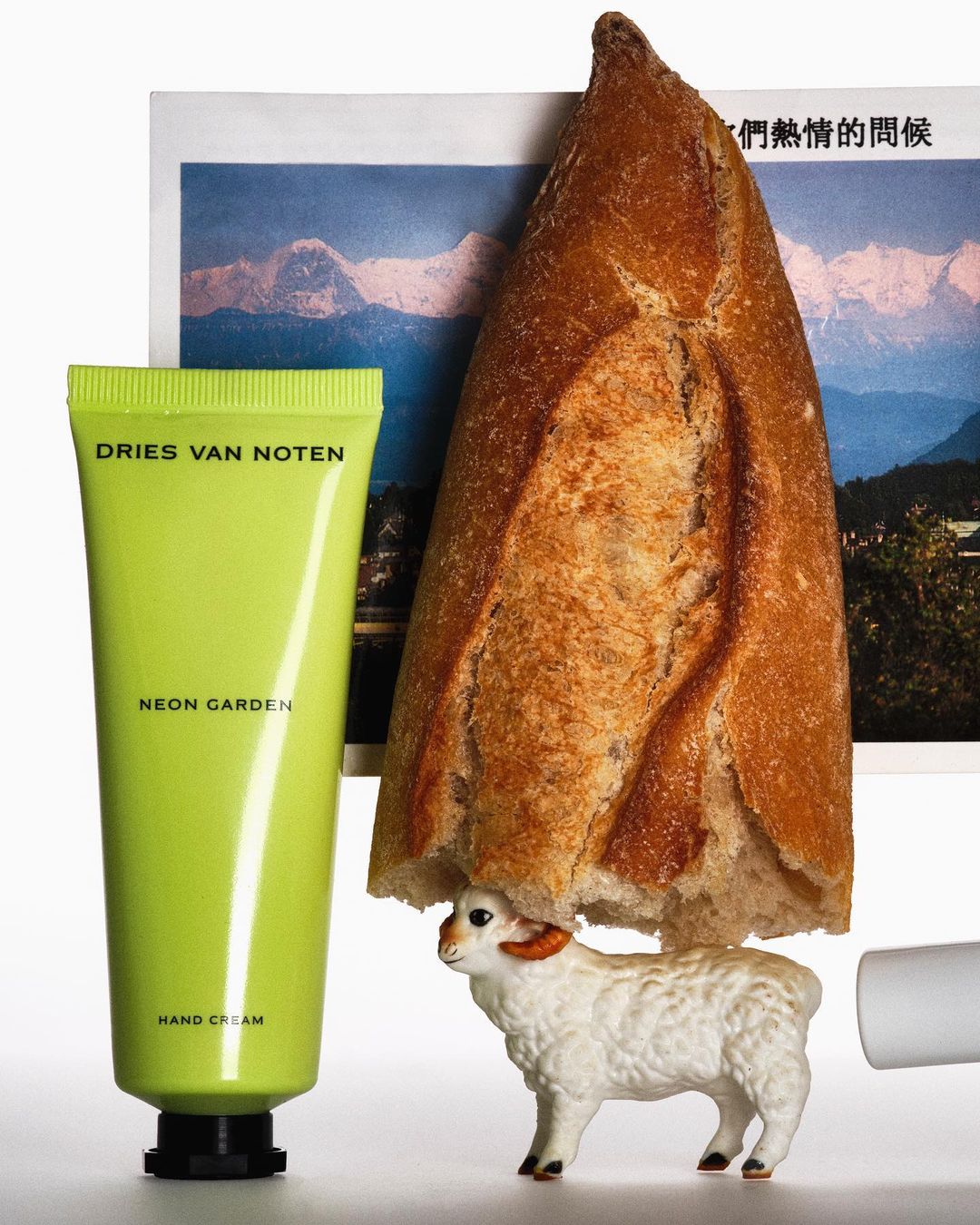 Still life featuring a neon-green hand cream tube, loaf of bread, toy sheep figurine, and a photo of snowy mountains on a white background.