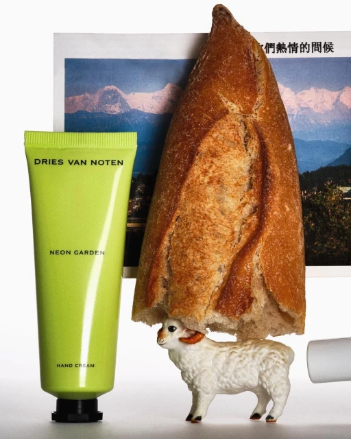 Still life featuring a neon-green hand cream tube, loaf of bread, toy sheep figurine, and a photo of snowy mountains on a white background.