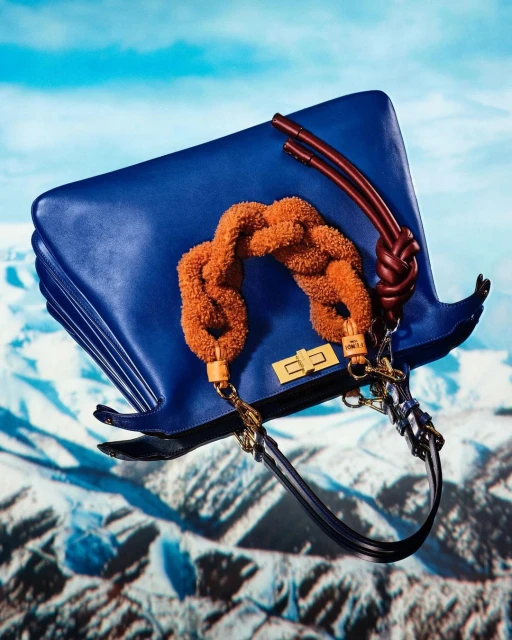 Blue leather tote bag with brown braided handle and straps, against a blurred mountain range and sky background.
