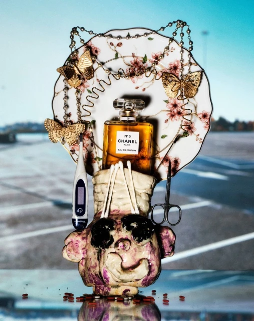 Still life with a Chanel N°5 perfume bottle, ceramic head sculpture, thermometer, scissors, plate with butterfly decor, against an urban backdrop.