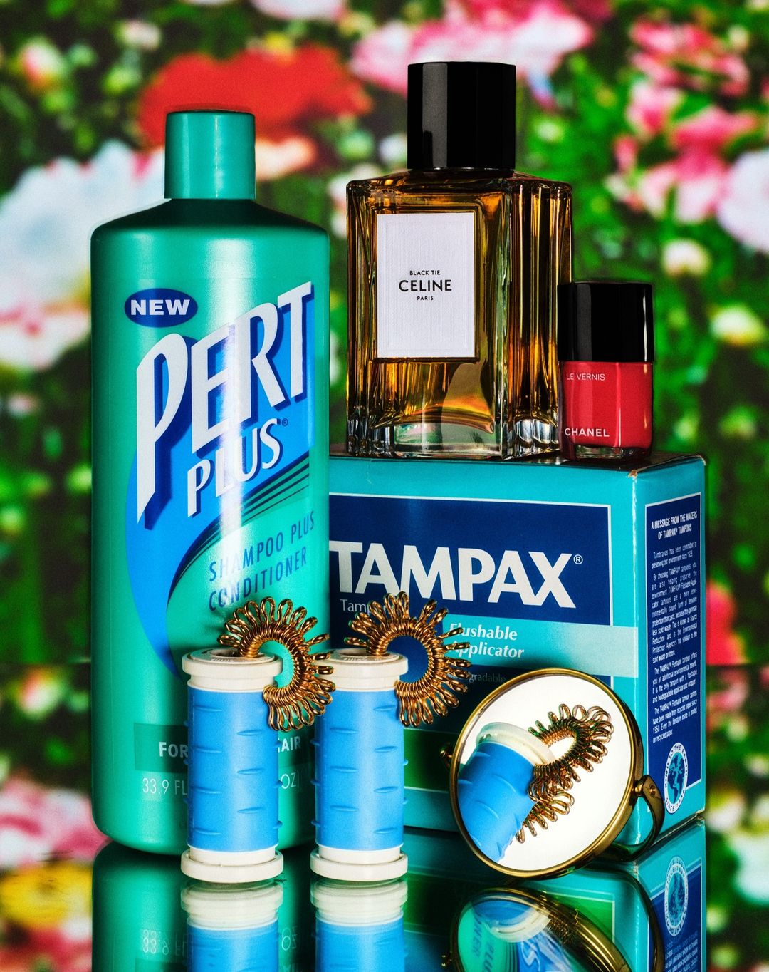 Still life featuring Pert Plus shampoo, Celine perfume, Chanel nail polish, Tampax box, hair rollers, and earrings against floral backdrop.