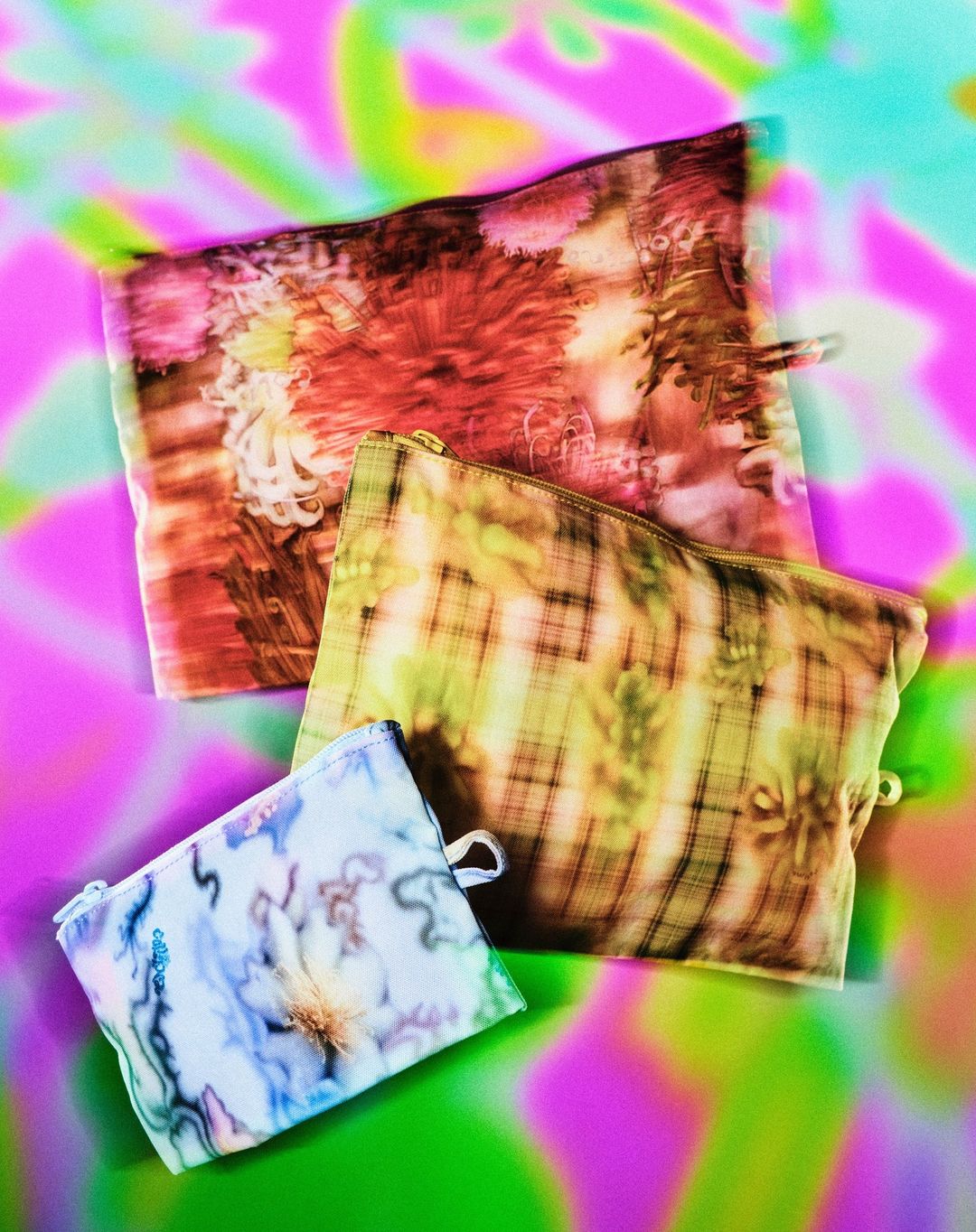 Three patterned zippered pouches with varying floral and geometric designs are arranged on a vivid green and pink abstract background.