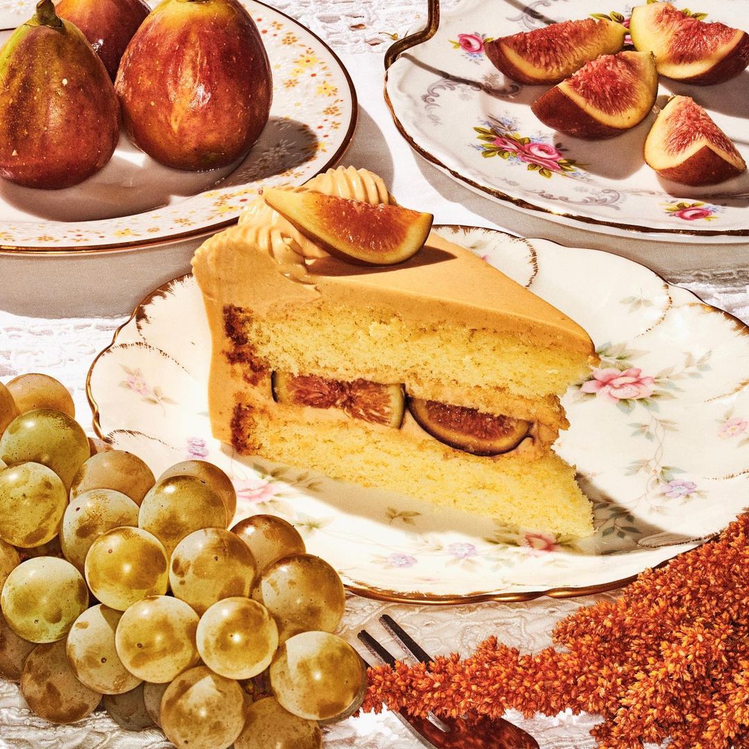 Close-up shot of a slice of fig cake served on a floral china plate, presented alongside figs and grapes on a white lace tablecloth.